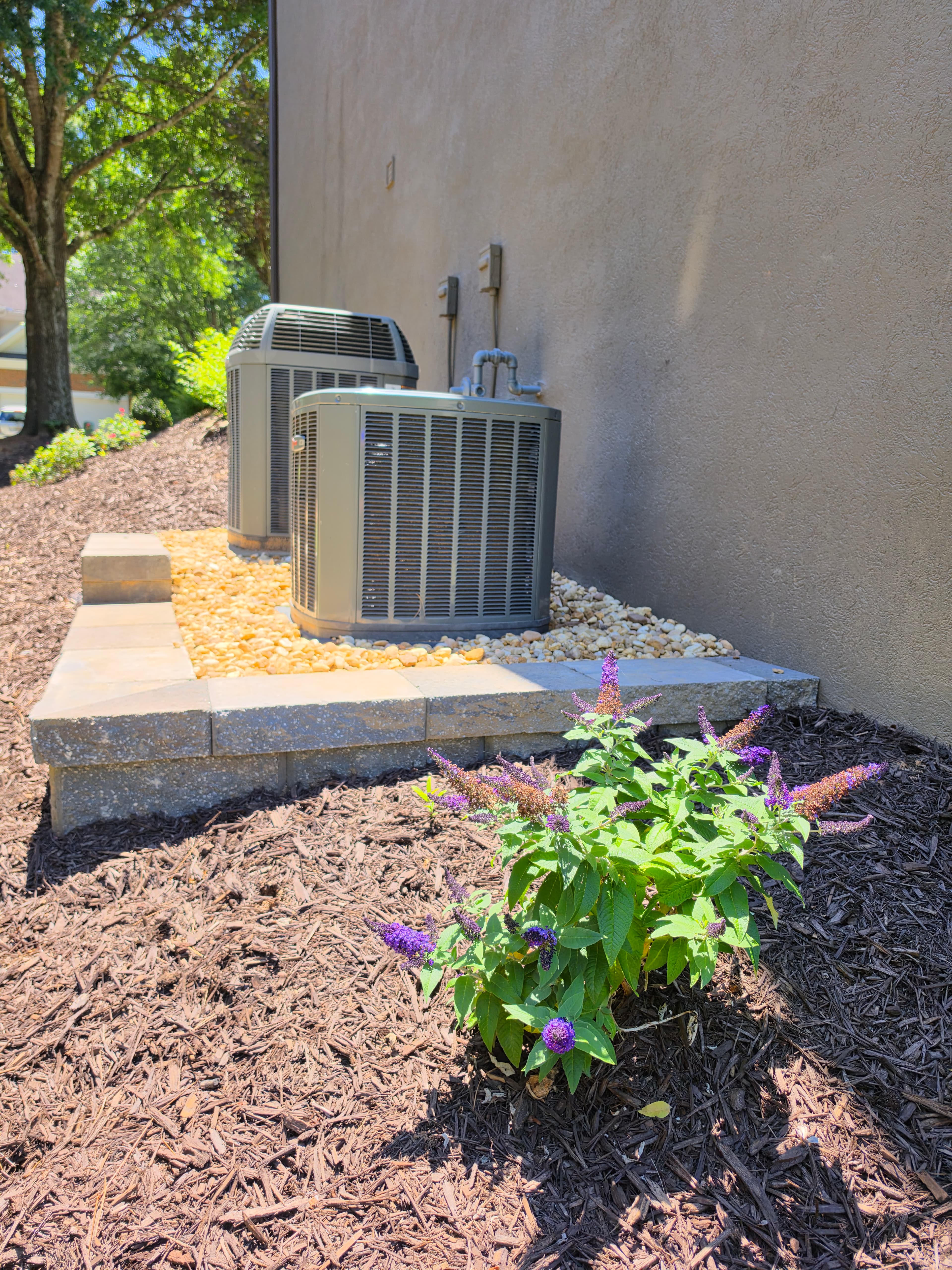 Two air conditioning units on a stone base next to a purple butterfly bush.