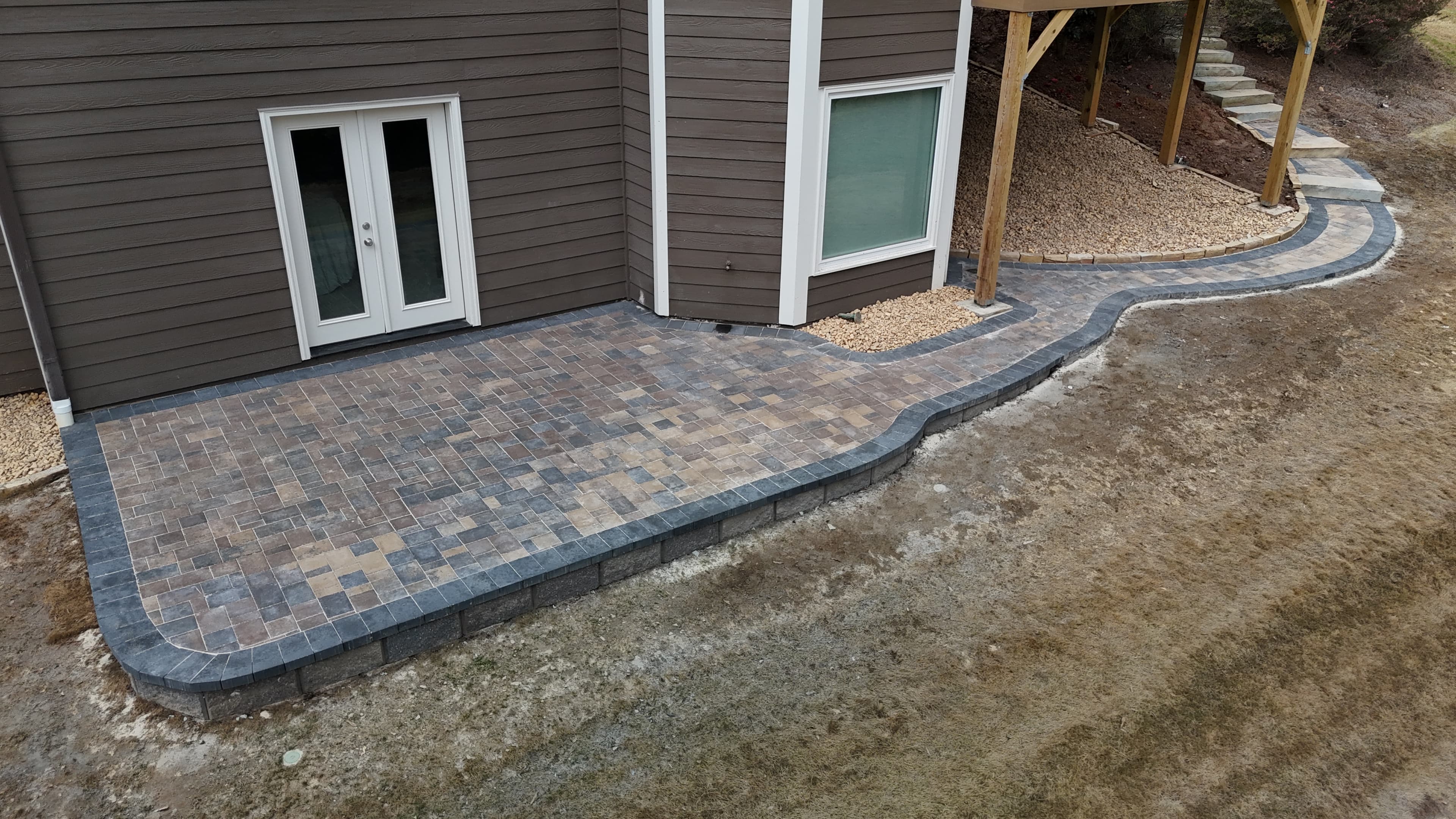 Stone paver patio and curved walkway bordering a brown house with white trim.