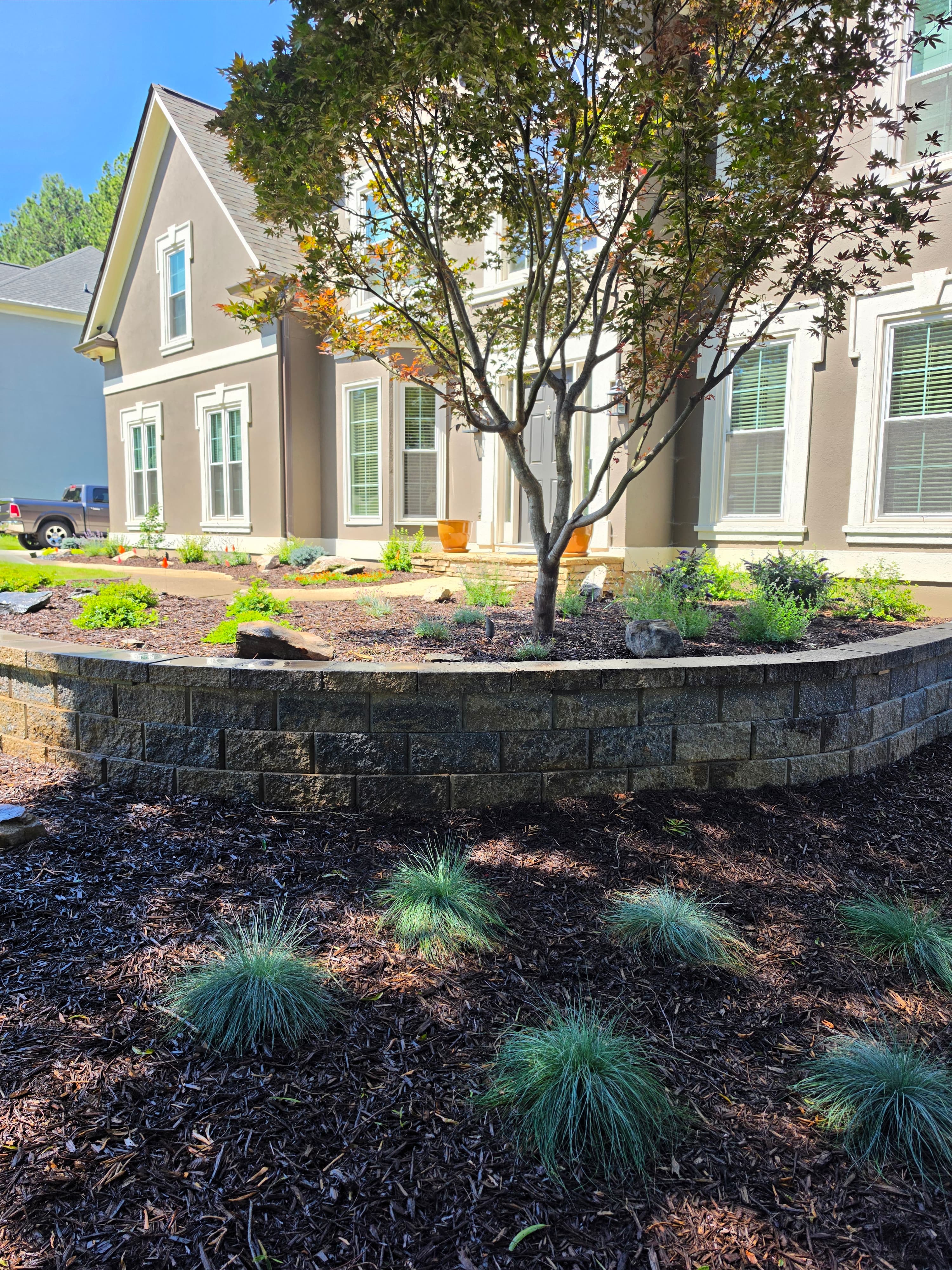 Two-story brown house with white trim behind a stone retaining wall and landscaped yard.