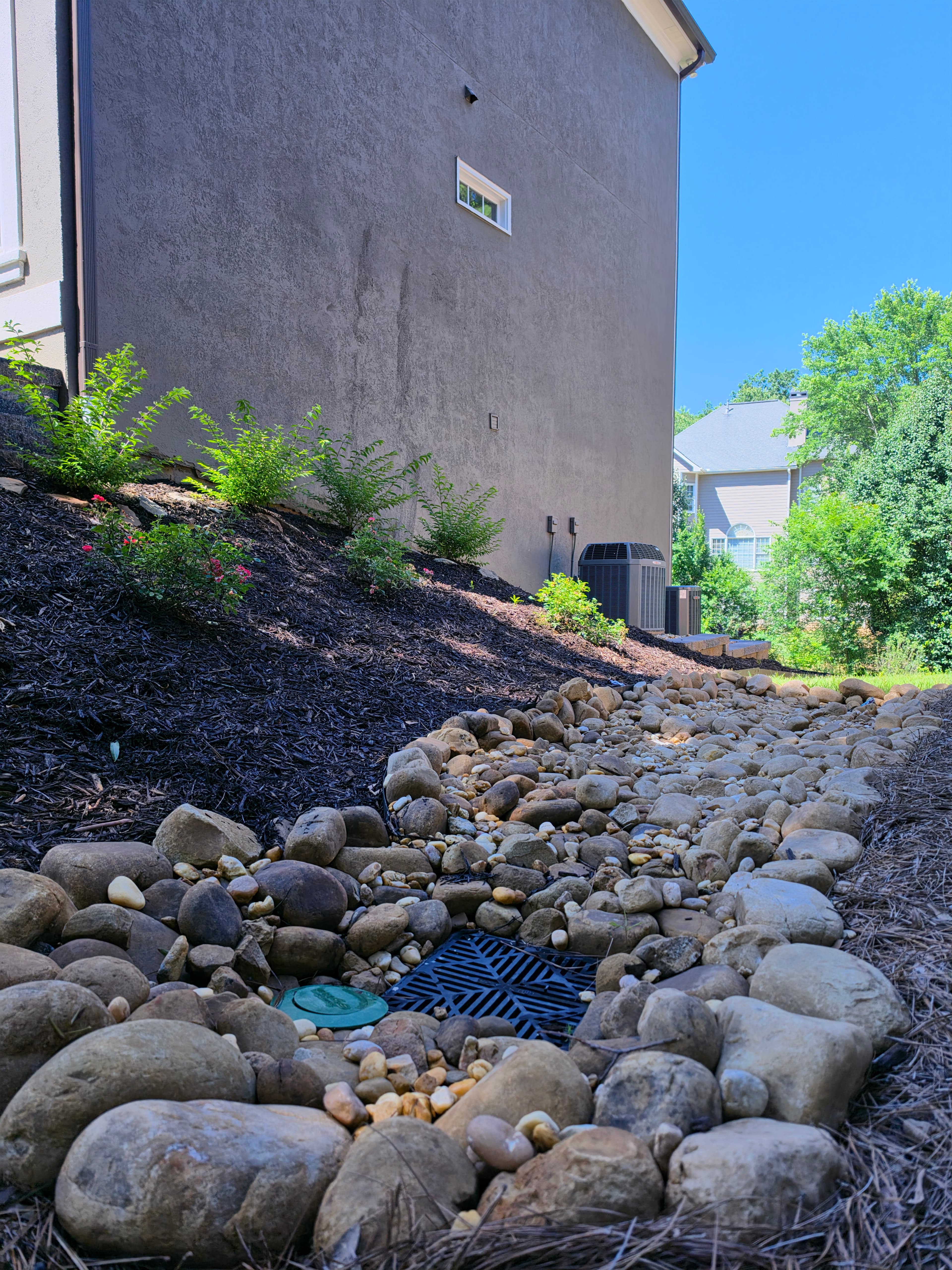 River rock dry creek bed with a drainage grate along a sloped, mulched garden.