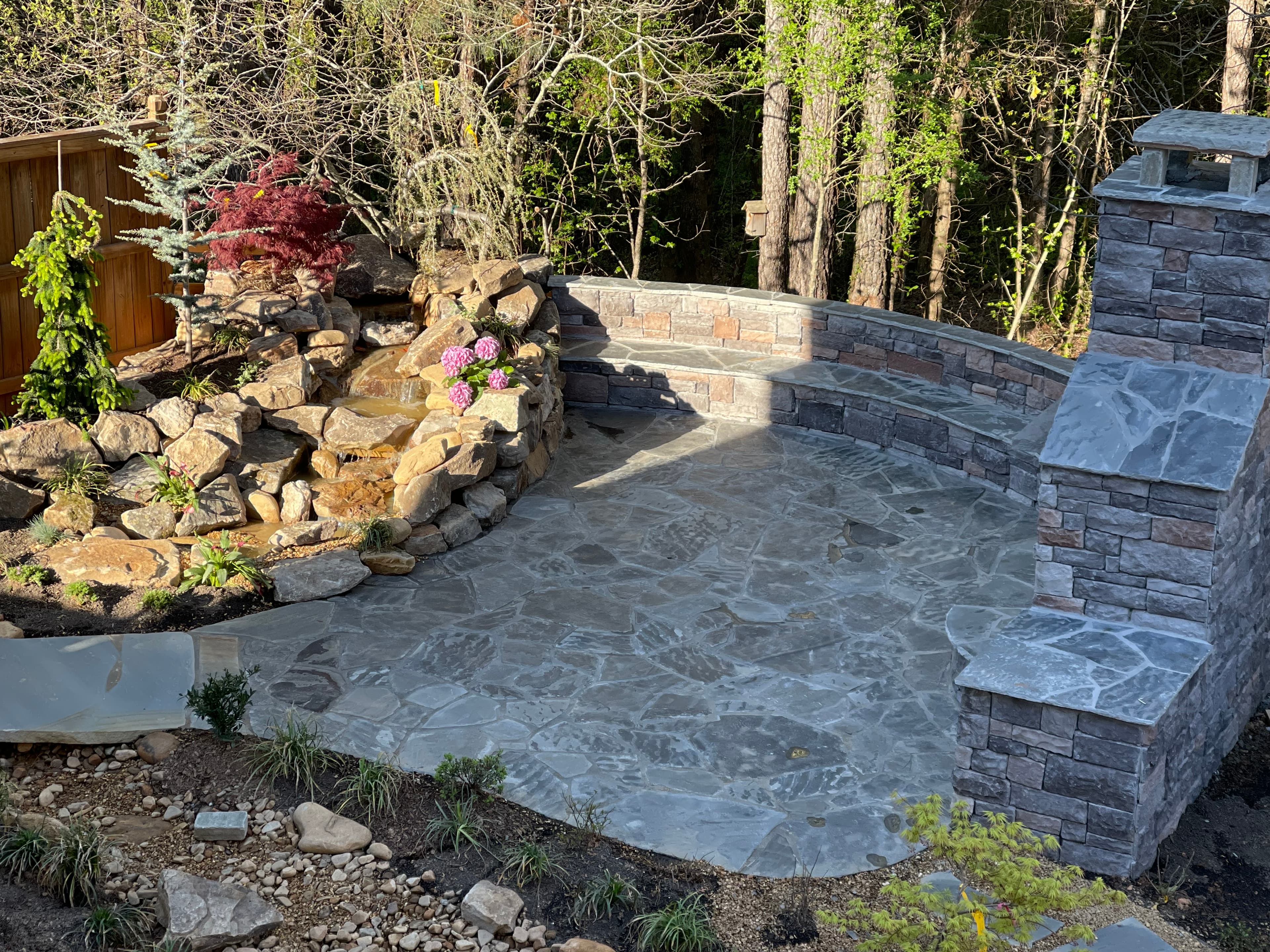 Flagstone patio with a curved stone bench, outdoor fireplace, and a tiered rock waterfall feature.
