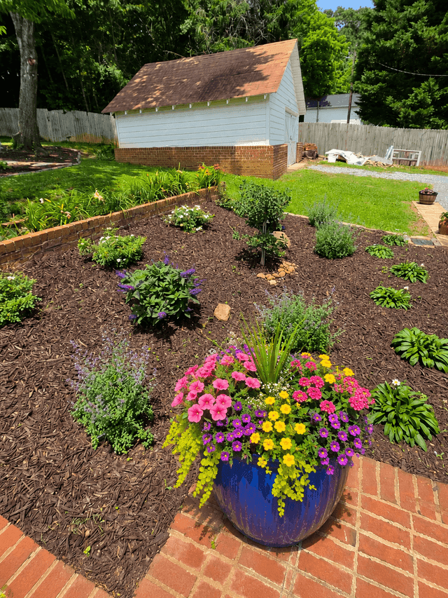 Vibrant blue planter with colorful flowers on a brick patio in a lush backyard garden.