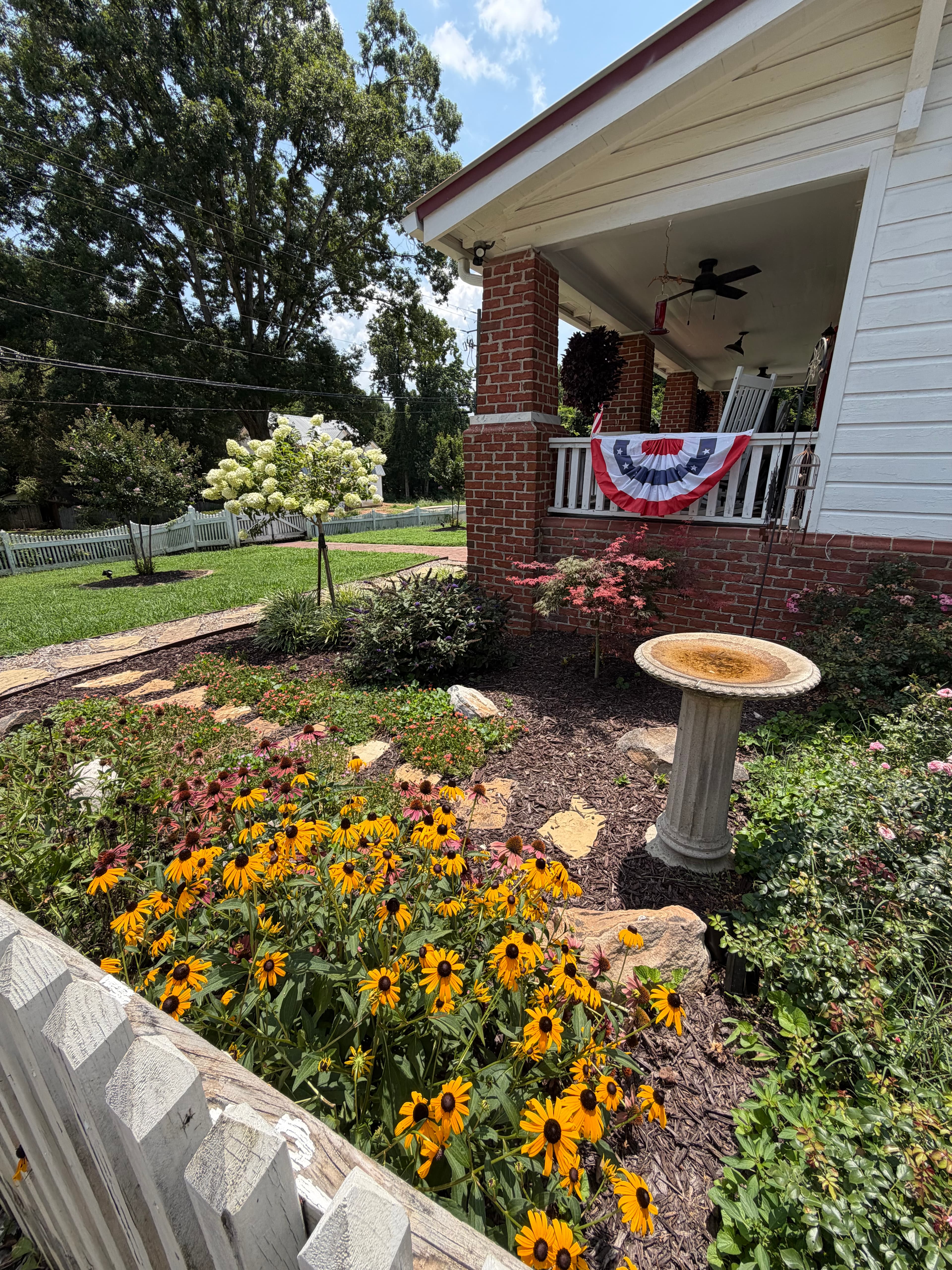 Yellow flowers and a stone birdbath in a garden beside a house with patriotic bunting.