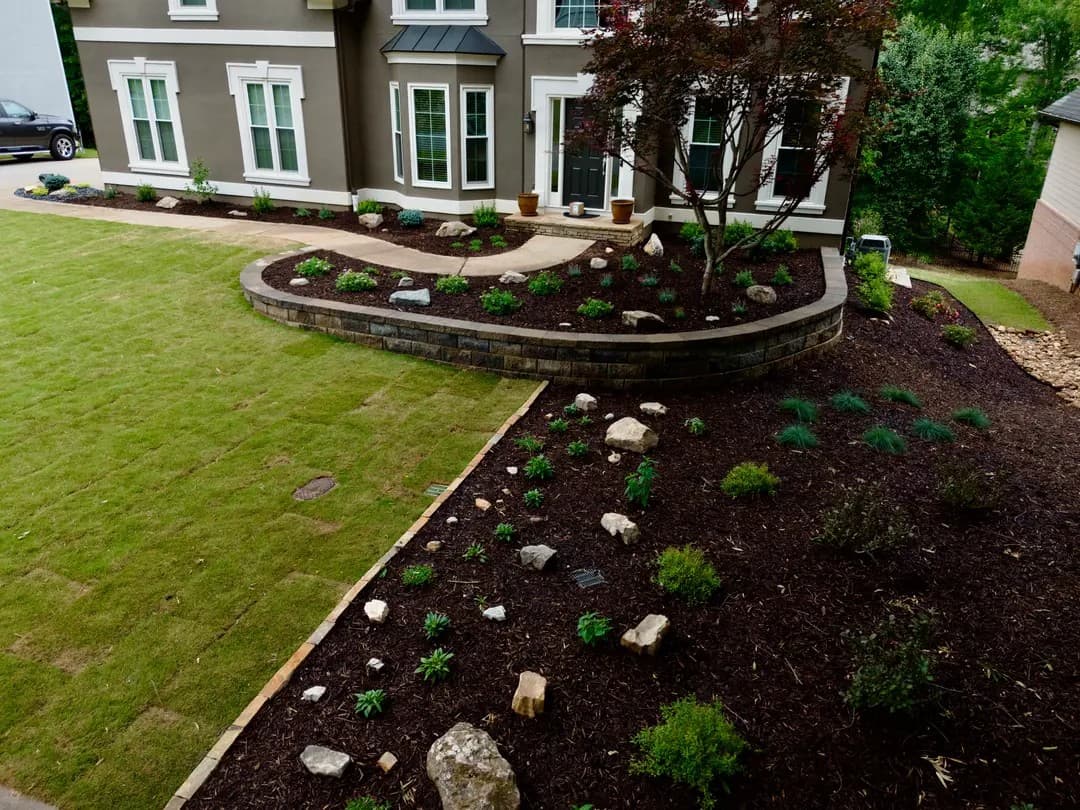 Landscaped front yard featuring a curved stone retaining wall, dark mulch, and new plants.
